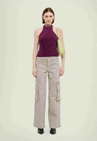 Sieviete, tērpusies bez piedurknēm purpursarkanā haltera topā, bēšās krāsas kravas biksēs, melnās kurpēs un turot gaismas zaļas plecu somu, stāv uz vienkārša fona.