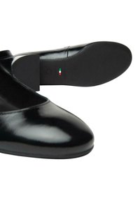 Scarpe in pelle nera con finitura lucida, punta arrotondata e tacco a blocco corto. La suola presenta una presa testurizzata e un'etichetta verde-rossa.