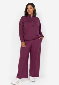 Chándal morado con parte superior de cremallera de manga larga y pantalones de pierna ancha. Tejido suave con puños acanalados; combinado con zapatillas blancas y grises.