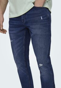 Mørkeblå denimjeans med en let slidfinish. Har en slim fit og diskret fading, med synlige sømme og et lille hul.