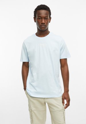 Junger Mann, der ein hellblaues s.Oliver Co. T-Shirt und beige Cargohosen trägt, steht mit einer Hand in der Tasche vor neutralem Hintergrund.