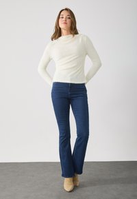 Top bianco a maniche lunghe con texture e jeans a zampa d'elefante blu scuro, abbinati a stivaletti beige su uno sfondo neutro.