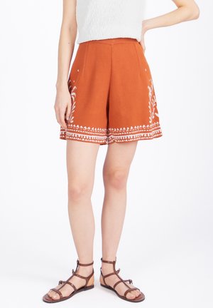 Naine, kes kannab roostepunaseid lühikesi pükse valge lillemustriga ja pruune rihmikuid tasaseid sandaale, seisab ühe käega puusal.