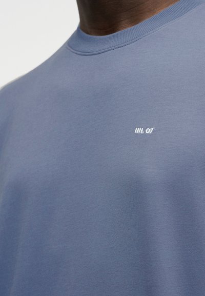 Κοντινό πλάνο ενός ατόμου που φοράει μπλε μπλούζα με στρογγυλή λαιμόκοψη και μικρό λευκό κείμενο "NN.07" στο στήθος.
