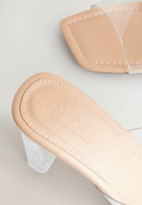 Sandales à talon transparent avec une semelle intérieure beige teintée, coutures blanches et détails de logo en relief. Design de talon bloc transparent.