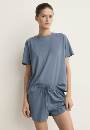 Kvinde står mod en ensfarvet væg iført en løs blå T-shirt med korte ærmer og matchende shorts med snøre.