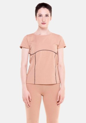 Jeune personne aux cheveux courts portant un haut beige à manches courtes et un pantalon ajusté assorti, debout devant un fond uni.