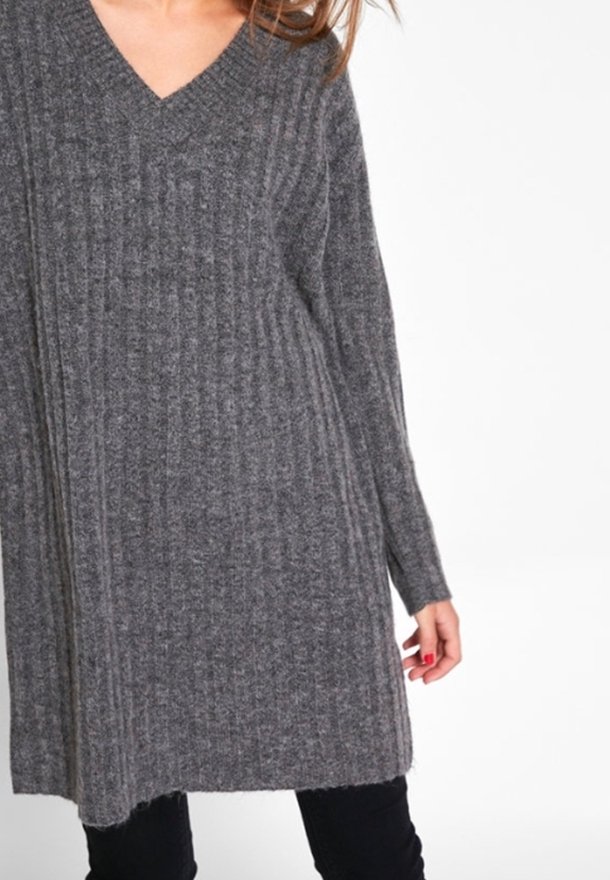 Žena v šedých žebrovaných pletených šatech s dlouhými rukávy a výstřihem do V, oblečená nad černými kalhotami, na bílém pozadí.