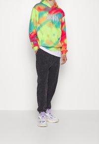 Mehrfarbige Batik-Hoodie in Grün, Gelb, Orange und Pink, kombiniert mit schwarzen strukturierten Jogginghosen und weißen Sneakern mit lila Akzenten.