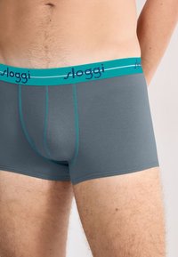 Gray boxer shorts with a teal elastic waistband featuring "sloggi" in dark blue. Smooth material and fitted design with subtle stitching details.