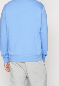 Sudadera azul claro con cuello redondo y mangas largas, con dobladillos y puños acanalados. Combinada con pantalones de chándal grises.