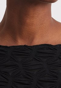 Schwarzes Top mit strukturiertem Design und gerüschten Paneelen. Der Halsausschnitt ist gerade und hebt das komplizierte Muster des Stoffs hervor.