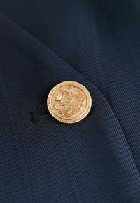 Arany díszgomb, domború mintával, amely egy texturált sötétkék szöveten helyezkedik el, ferde bordázattal és egy kis cérna hurokkal.