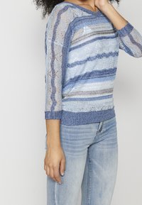 Pull tricoté bleu clair avec des rayures verticales de différentes nuances, doté d'un motif en dentelle transparent et de manches trois-quarts. Poignets texturés.