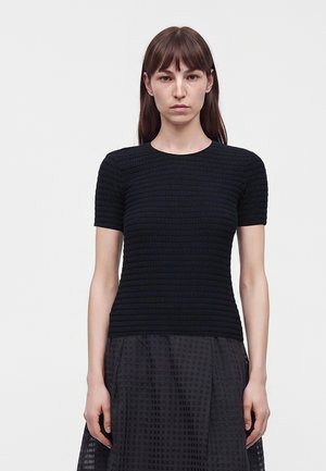 Femme aux cheveux bruns et raides porte un haut noir texturé moulant à manches courtes et une jupe noire à carreaux, debout devant un fond uni.