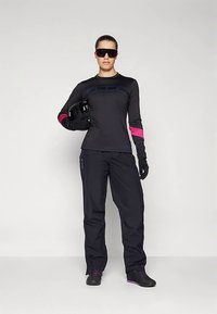Schwarzes Langarm-Top mit blauen Akzenten, pinken Armbändern und schwarzer Hose, ergänzt durch schwarze Handschuhe und Sonnenbrille, hält einen Helm.