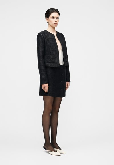 Čierny tweedový kabátik s krátkym strihom a dvoma prednými vreckami, v kombinácii so zodpovedajúcou sukňou s gombíkmi, priesvitným topom a špicatými topánkami.