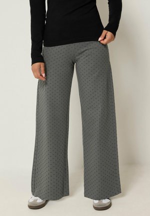 Persona que lleva pantalones grises de pierna ancha con pequeños lunares, camiseta negra de manga larga y zapatillas blancas, de pie contra un fondo liso.
