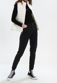 Bílá prošívaná vesta přes černé triko s dlouhým rukávem, doplněná černými cargo kalhotami a černými teniskami Converse s vysok ým svrškem. Jednoduchý, funkční design.