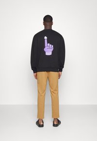 Mann in einem schwarzen Sweatshirt mit lila grafischer Hand und beigen Hosen, der vor einer schlichten weißen Wand steht.