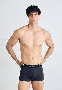 Schwarze Boxershorts mit einem Logo-Bund, aus einem glatten, dehnbaren Material, angepasst und in Länge bis zur Mitte des Oberschenkels, Modell steht mit Händen auf den Hüften.