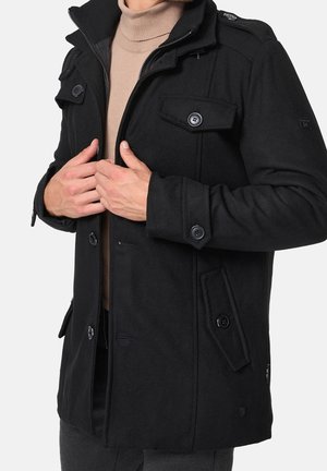 Mann trägt schwarzen, geknöpften Wollmantel über beigem Rollkragenpullover und richtet die Mantelvorderseite mit beiden Händen gerade.