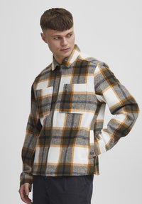 Fleece jacket featuring a large plaid pattern in shades of white, orange, brown, and gray. It has two chest pockets and a zip closure.