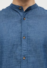 Κλείσιμο σε όψη ενός ατόμου που φοράει μπλε πουκάμισο από λινό με γιακά μανταρίνι και τέσσερα καφέ κουμπιά στη μπροστινή πλευρά.