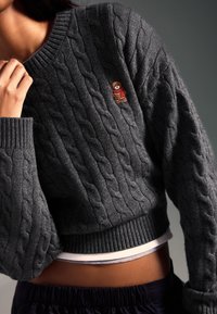 Siv pulover z debelo pletenim vzorcem, dolgimi rokavi, rebrastimi manšetami in spodnjim robom. Na prsih ima majhen našitek z medvedkom. Mehka tekstura, priložnostni kroj.