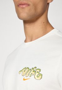 Camiseta de algodón blanca con un gráfico en verde y amarillo que dice "HIKE" y un logo de Nike en naranja. Textura suave, cuello redondo, manga corta.