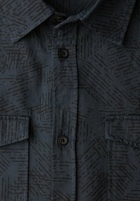 Ciemnoniebieska koszula z subtelnym czarnym geometrycznym wzorem, zapinana na guziki, z dwoma kieszeniami na piersi. Wykonana z teksturowanej tkaniny.