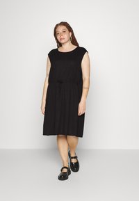 Vestido preto sem mangas com decote redondo e cintura com cordão. Textura suave, até ao joelho, combinado com sapatos pretos de tiras.
