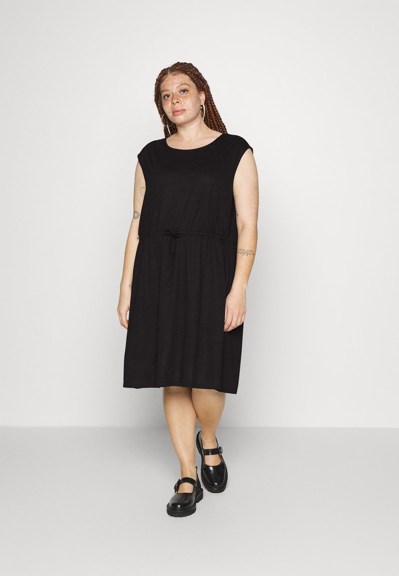 Vestido preto sem mangas com decote redondo e cintura com cordão. Textura suave, até ao joelho, combinado com sapatos pretos de tiras.