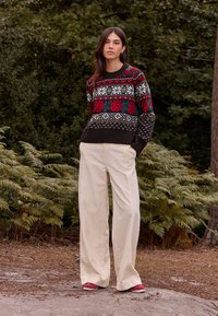 Pull en tricot avec motif floral rouge et vert, ourlet côtelé noir, associé à un pantalon crème à jambes larges et des chaussures rouges. Fond naturel.