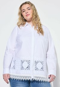Bílá košile s knoflíky, zdobená texturovanou krajkou, obsahující vyšívané květinové detaily a čtvercové krajkové výřezy na lemu. Casual střih s dlouhými rukávy.