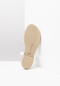 Sandale blanche avec une semelle en caoutchouc beige, présentant un motif texturé, des brides ajustables autour de la cheville et une étiquette de taille indiquant "37".