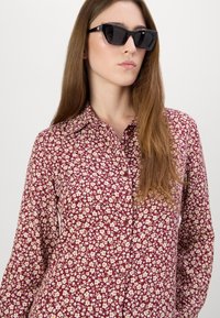 Bluză butonată vânătă cu un mic model floral în galben și alb; are guler și mâneci lungi. Modelul poartă ochelari de soare negri cu formă de ochi de pisică.