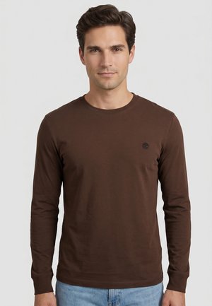 Homme aux cheveux courts et bruns portant un t-shirt marron à manches longues et un jean bleu clair, debout devant un fond uni gris clair.