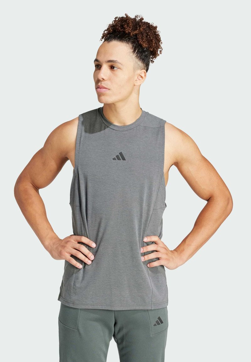 T-shirt de sport gris sans manches avec un col rond et une texture douce, arborant un petit logo noir sur la poitrine. Associé à un pantalon vert olive.