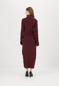 Burgunderfarbenes Langarm-Strickkleid mit hohem Kragen, seitlichen Schlitzen und Knopfakzenten an den Ärmeln. Dazu werden schwarze High Heels getragen.