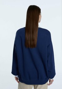 Granatowy, oversize'owy sweter z długimi rękawami, ściśle zwężanym mankietem i wykończeniem. Gładka faktura, minimalistyczne detale oraz luźny krój, noszony na koszuli z kołnierzykiem.