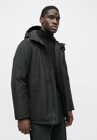 Giacca imbottita di color verde scuro con zip frontale, cappuccio e tasche laterali. Texture liscia, vestibilità rilassata e design minimalista.