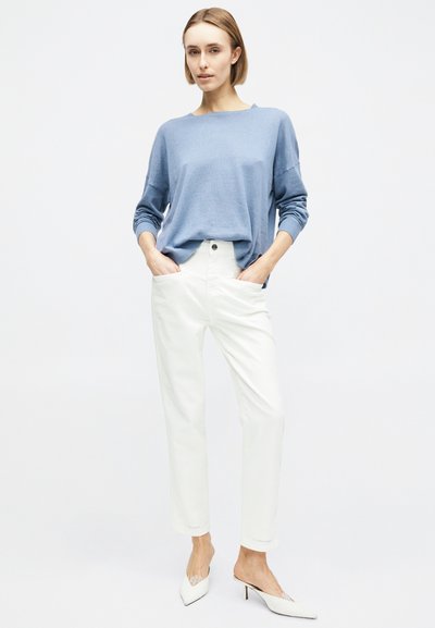 Γυναίκα με κοντά μαλλιά στέκεται με τα χέρια στις τσέπες, φορώντας ανοιχτό μπλε μακρυμάνικη μπλούζα, λευκό παντελόνι ψηλόμεσο και λευκές μυτερές γόβες.