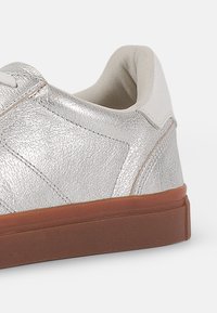 Silver leather sneaker with a textured finish, white interior lining, and a brown rubber sole. Features detailed stitching and smooth accents.