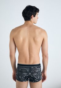 Crni bokserice s uzorkom, s paisley dizajnom, koje imaju kontrastni pojas. Model je prikazan s leđa bez vidljivog gornjeg dijela.