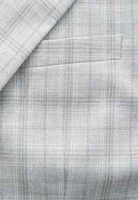 Tela gris claro con un patrón de cuadros que presenta finas líneas en marrón y blanco. Contiene un único detalle de bolsillo. Textura suave, material tejido.