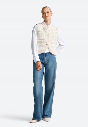 Gilet bianco con texture a motivo quadrato sopra una camicia bianca a maniche lunghe, abbinato a jeans a gamba larga azzurri e scarpe beige.