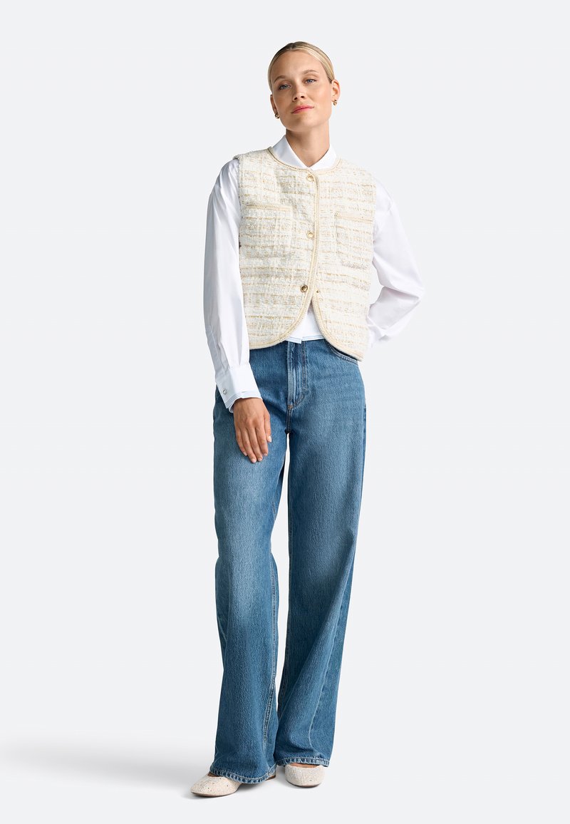 Gilet bianco con texture a motivo quadrato sopra una camicia bianca a maniche lunghe, abbinato a jeans a gamba larga azzurri e scarpe beige.