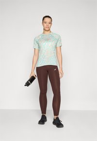 Camiseta atlética azul claro con remolinos, combinada con leggings marrones y zapatillas negras. La modelo sostiene una botella de agua negra. Diseño simple y ajustado.
