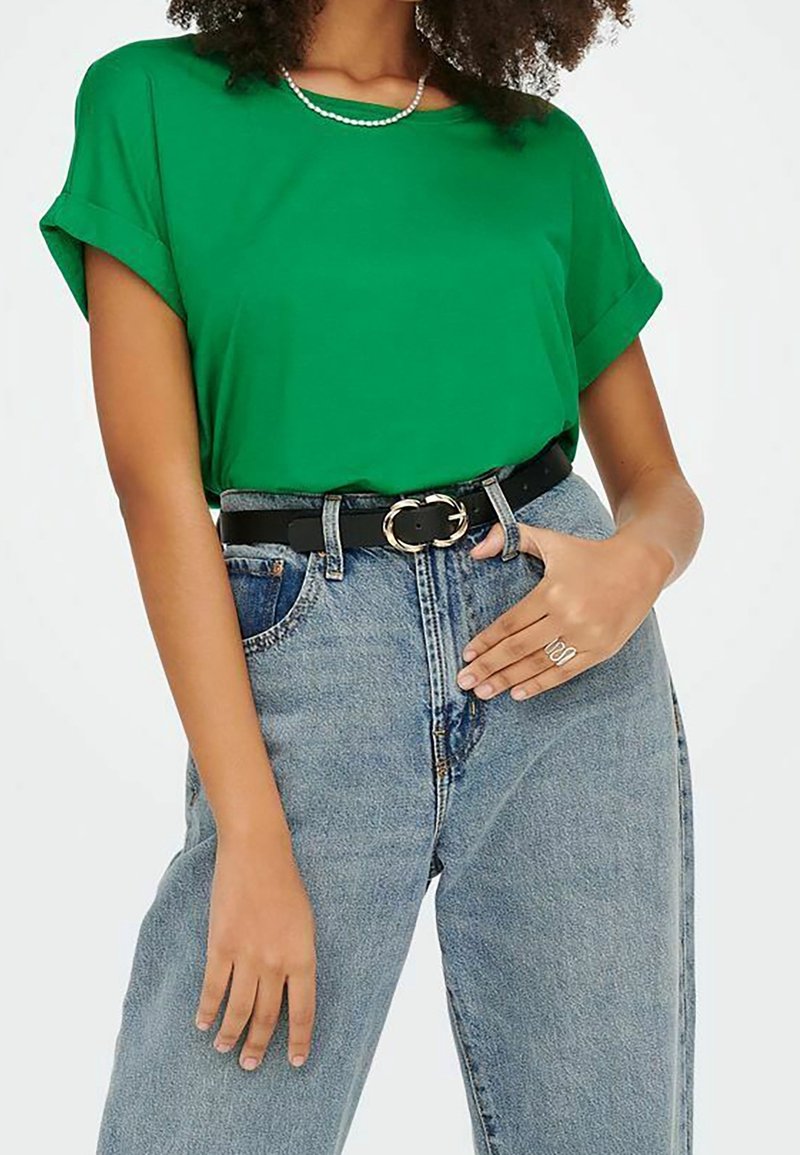 Femme portant un haut vert à manches courtes, un jean bleu clair taille haute avec ceinture noire, un collier de perles et une bague double en argent à la main droite.